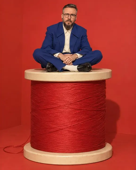 A bearded man in a cobalt suit sits cross-legged on top of a giant spool of red thread taller than a person