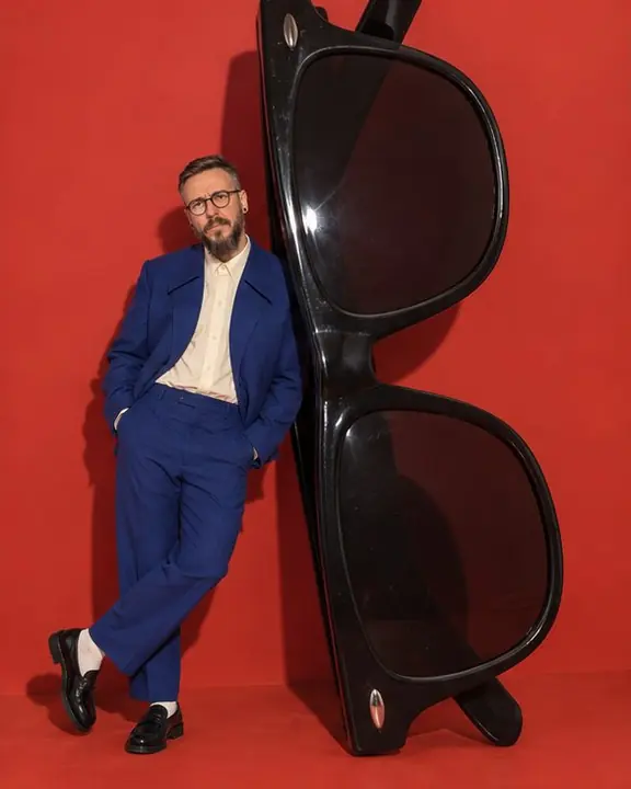 A man leans against a pair of giant black sunglasses standing upright, three times his height, on a red set