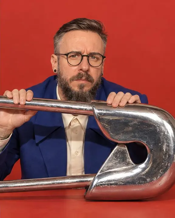 Half-length of a bearded man in round glasses peeking over a giant chrome safety pin lying across the frame