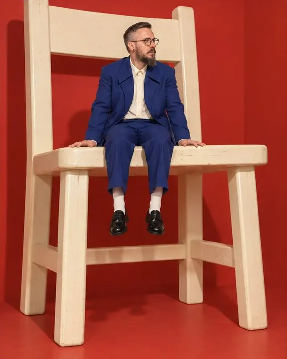 A man in a cobalt suit sits on a giant cream wooden chair, his legs dangling far above the floor