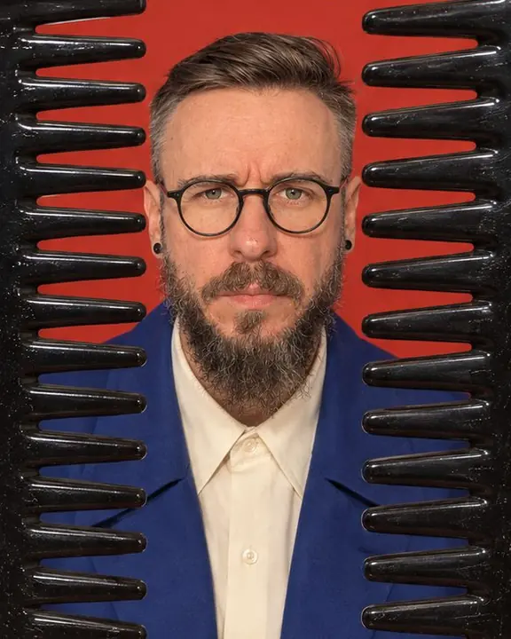 Head and shoulders of a bearded man framed between the teeth of a giant black comb standing upright