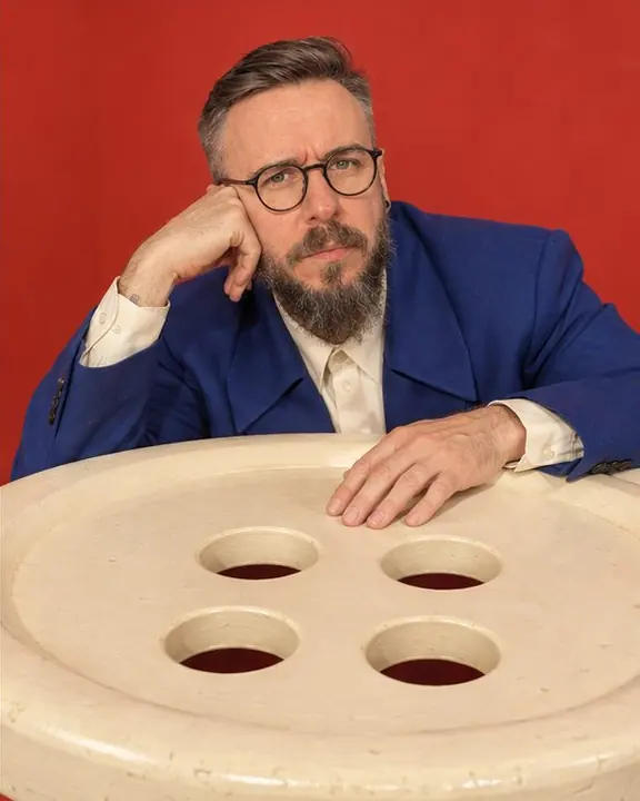 A hand with a cobalt suit cuff resting on a giant cream shirt button the size of a table