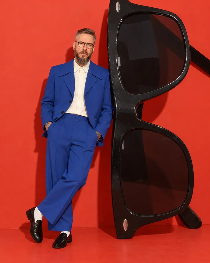 A man leans against a pair of giant black sunglasses standing upright, three times his height, on a red set