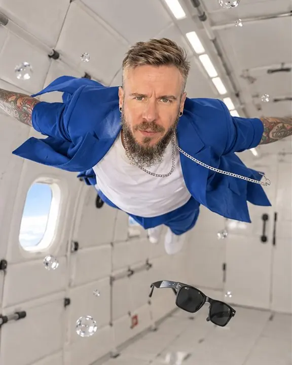 A bearded man floats horizontally across a white padded cabin, arms spread, his blue suit jacket billowing open