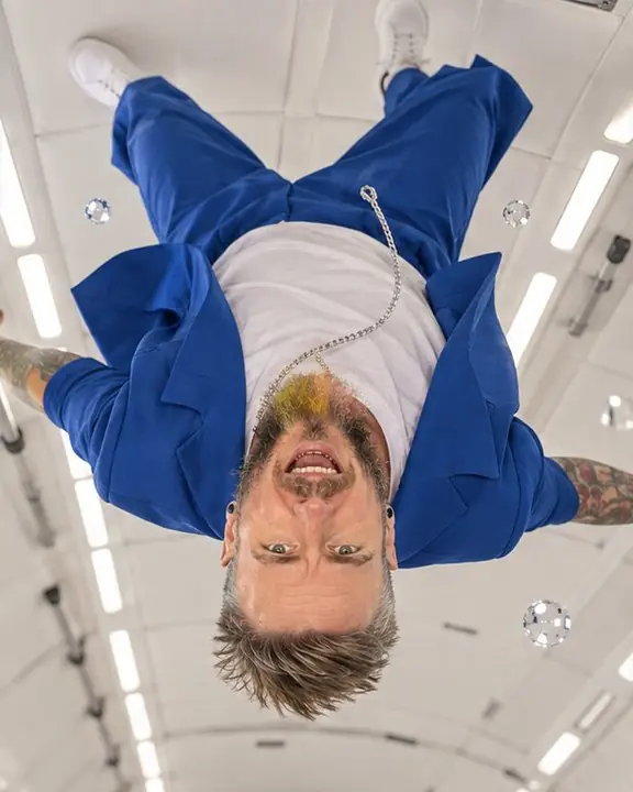 Upside down near the cabin ceiling, a bearded man in an electric blue suit laughs at the lens, hair standing up