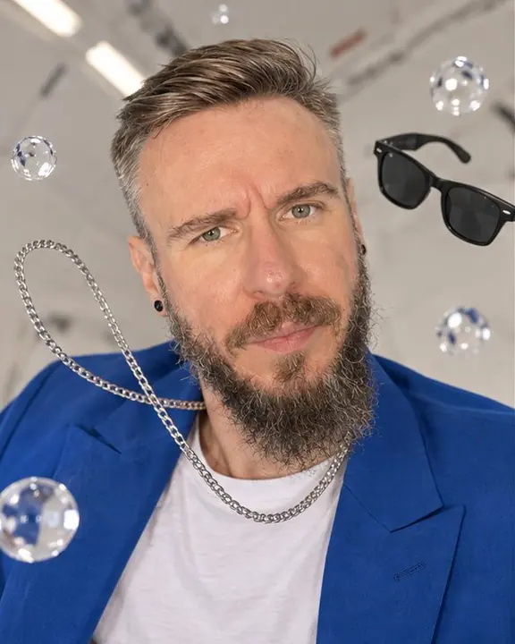 Head and shoulders of a bearded man as a silver chain and black sunglasses drift weightless around his face