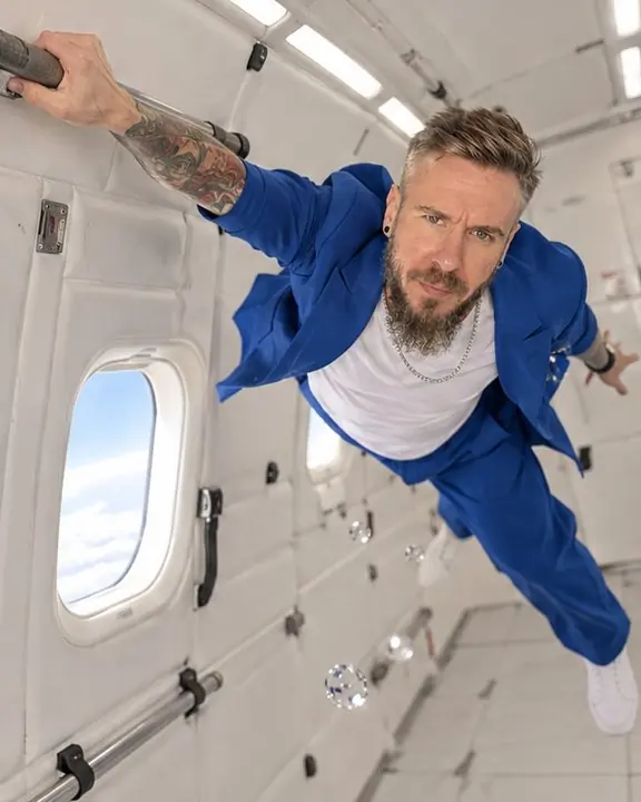 A man pushes off a handrail and stretches diagonally across the cabin, a small window of bright sky behind