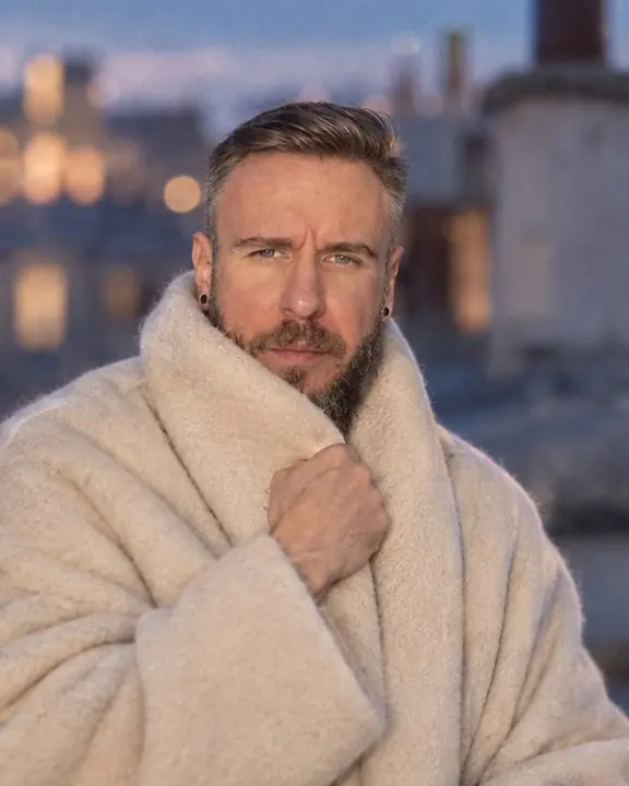 Waist-up shot of a man wrapped high in a cream cashmere blanket-coat, a soft diffusion glow on his face