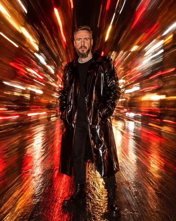A man in a glossy black vinyl coat stands in the middle of a burst of red and amber light lines